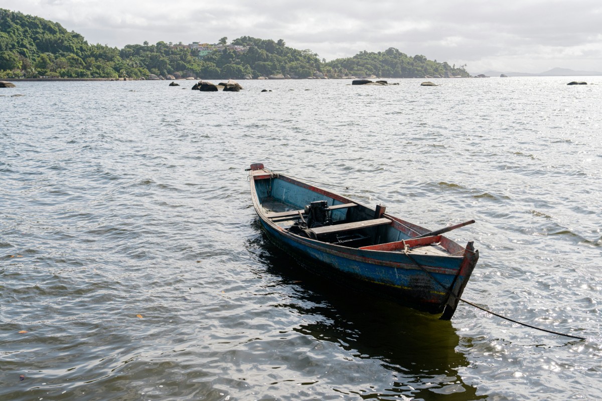 A small, empty wooden boat floats on calm water near a forested shoreline under a cloudy sky.