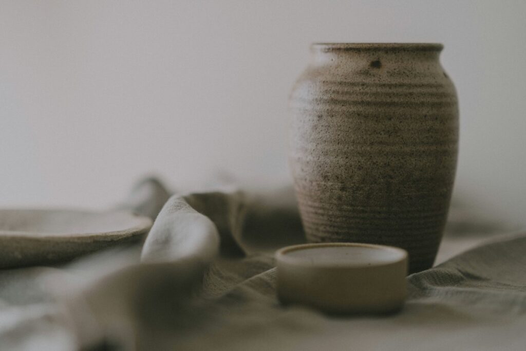 A textured, brown ceramic vase on a soft draped fabric, with a matching round lid beside it. Blurred background.