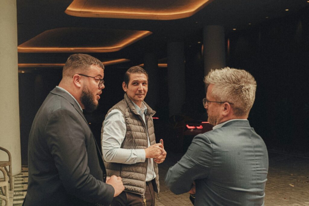 Three men converse in a dimly lit indoor space. The man in the middle wears a puffy vest, while the others wear jackets.