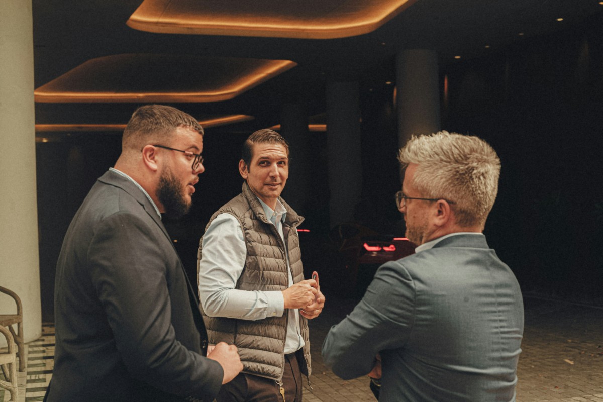 Three men converse in a dimly lit indoor space. The man in the middle wears a puffy vest, while the others wear jackets.