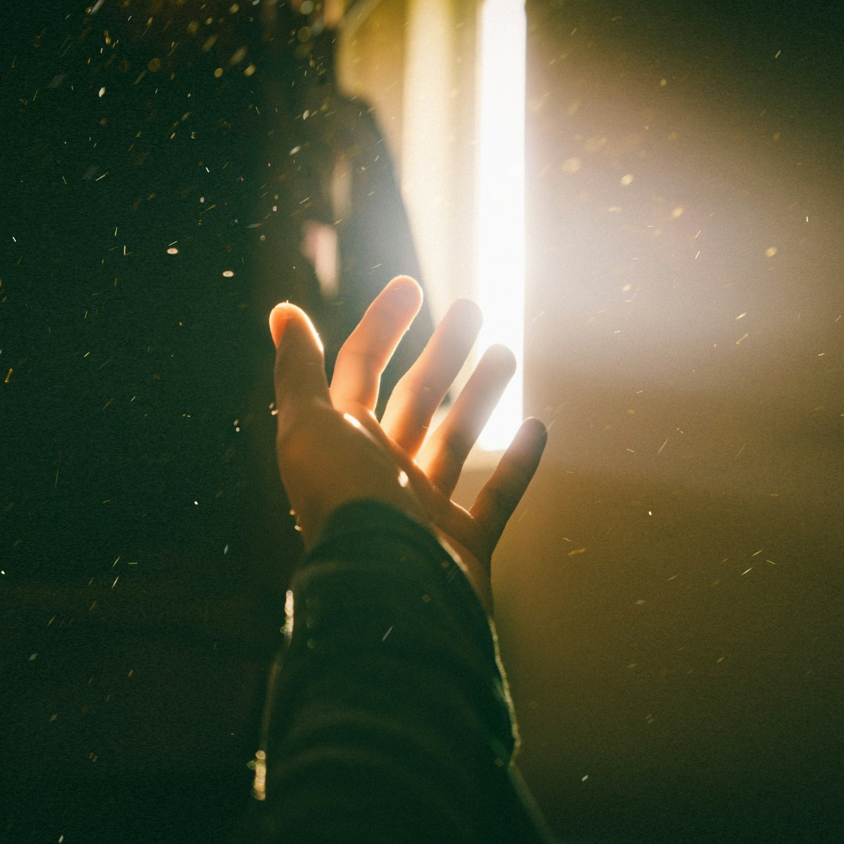 A hand reaching towards a bright light source, surrounded by particles in a dimly lit environment.