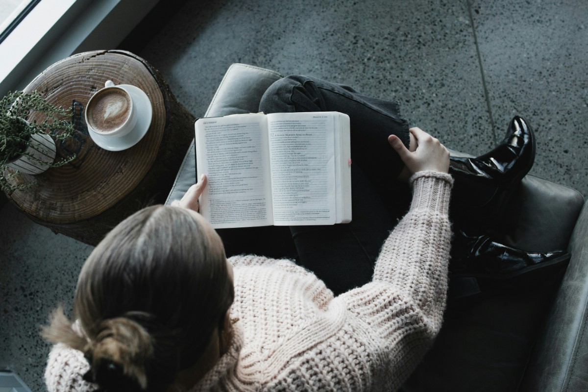Person in a beige sweater and black boots reading a book on a gray couch, with a cup of coffee on a small wooden table nearby.