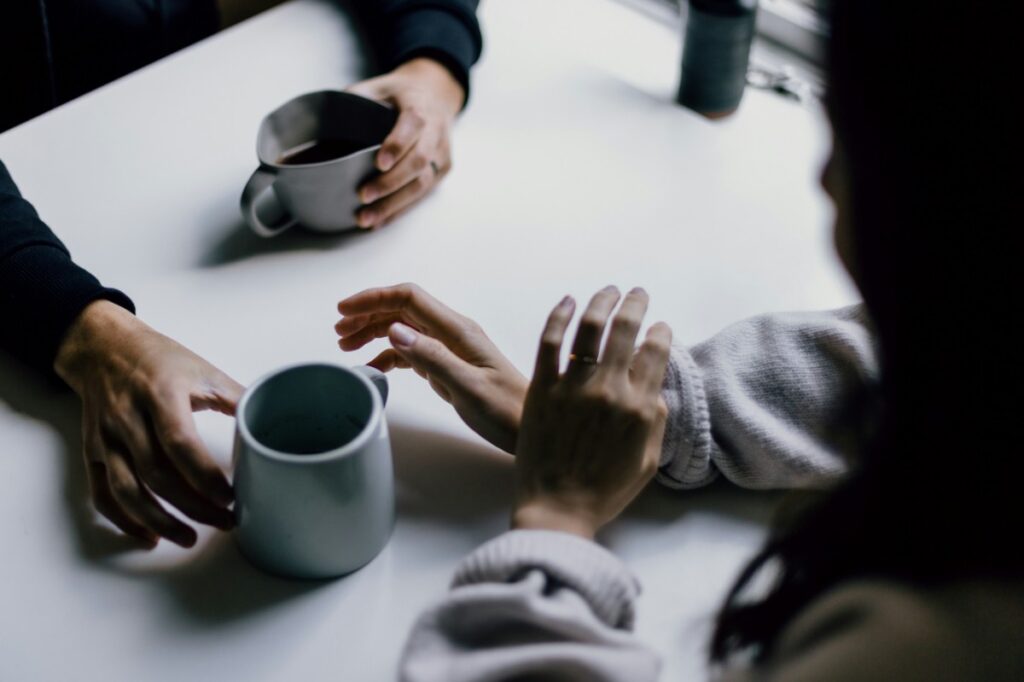 Two people sitting at a table with mugs, having a conversation. Their hands are visible, one reaching towards a mug.