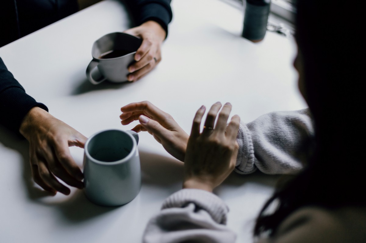 Two people sitting at a table with mugs, having a conversation. Their hands are visible, one reaching towards a mug.