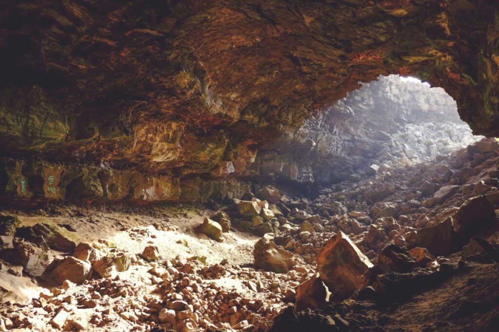 Sunlight illuminates the rocky interior of a cave with a rugged entrance, featuring scattered stones and earthy textures.