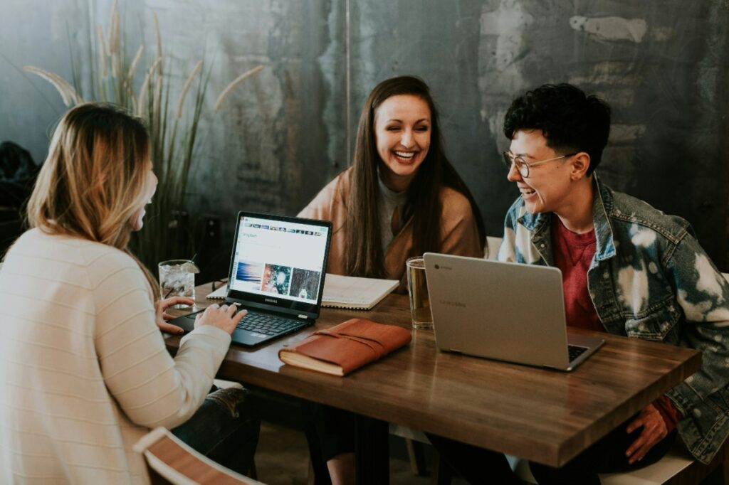 Three people are sitting at a wooden table with laptops, smiling and chatting. Books and a potted plant are on the table.