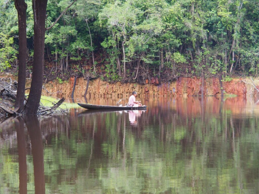 A person sits in a small wooden boat on a calm river with dense green forest and a red earth bank in the background.