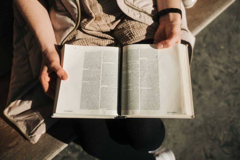 Person seated, holding and reading an open book with white pages and black text, in sunlight.