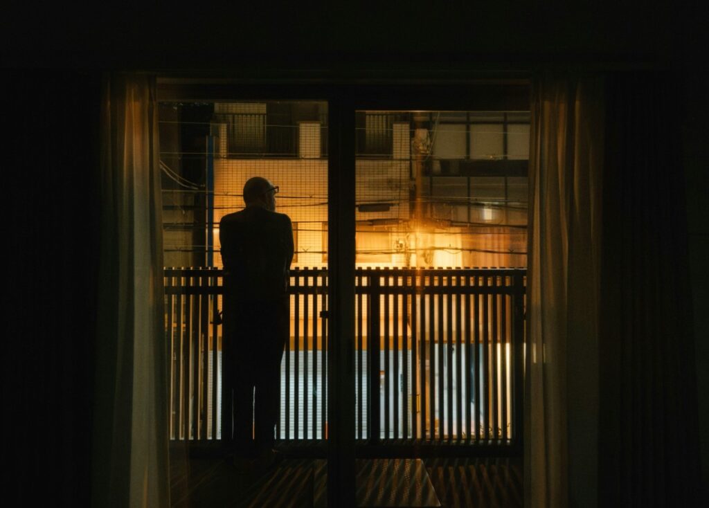 Silhouette of a person standing by a window, looking out at an illuminated urban scene at night.