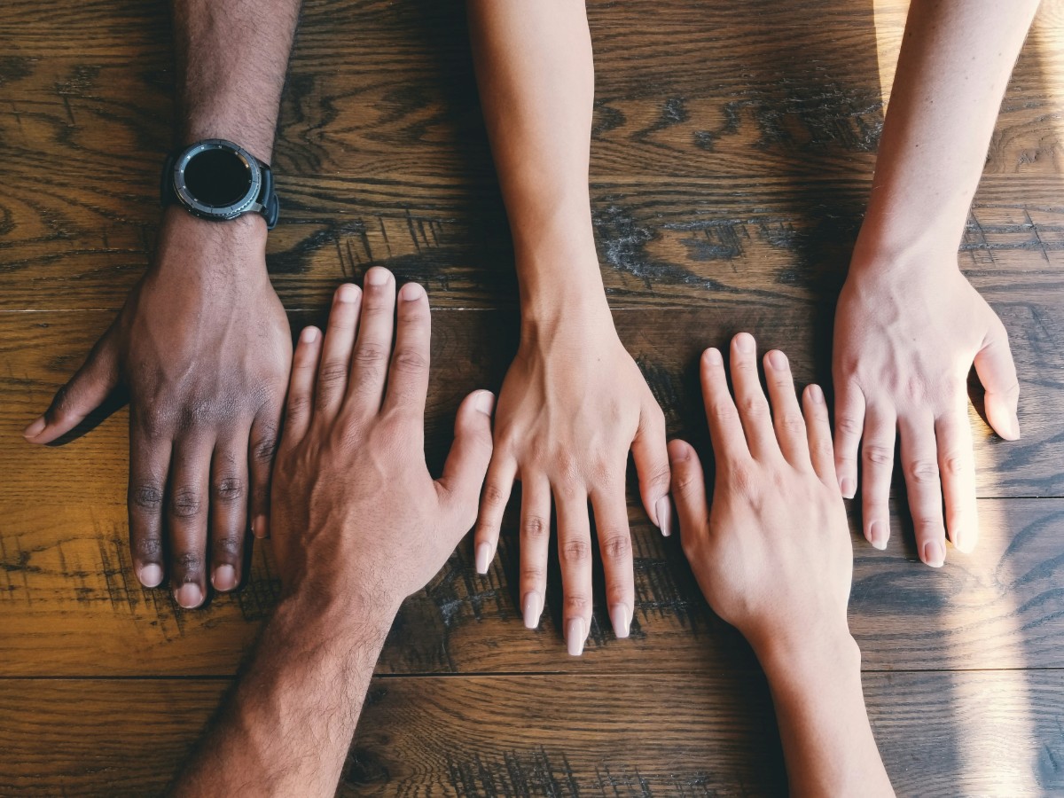 Four hands of different skin tones are laid flat on a wooden table, all reaching toward the center.