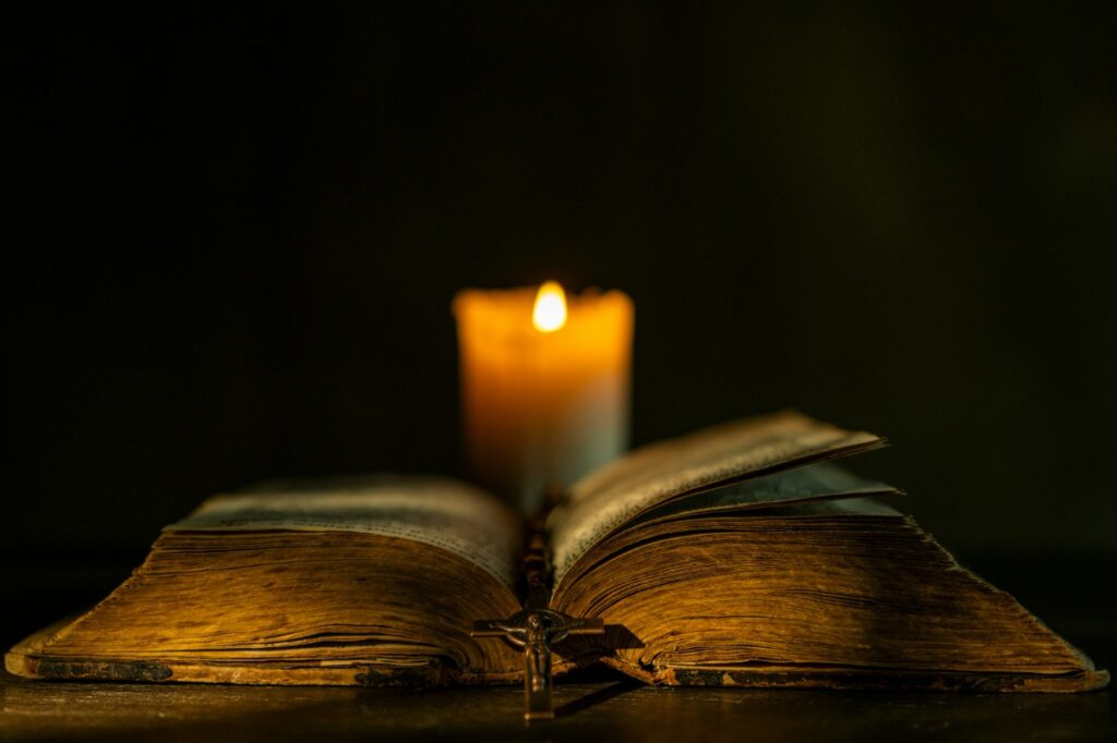 An open, antique book with yellowed pages lies on a table, illuminated by a burning candle in the background.