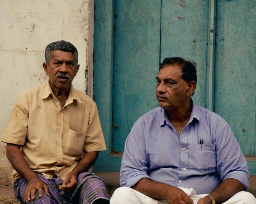 Two men sitting in front of a blue door, engaged in conversation. One wears a tan shirt and patterned pants, the other a light blue shirt and white pants.