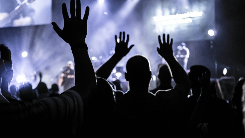 Audience with raised hands silhouetted against a bright concert stage backdrop.