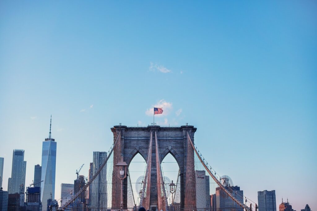 Brooklyn Bridge with a U.S. flag atop, set against a blue sky and the Manhattan skyline, including One World Trade Center.