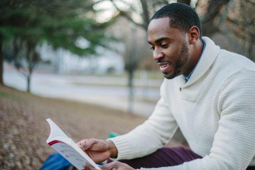 A man in a white sweater sits outdoors on a bench, reading a book. Trees and a path are visible in the background.