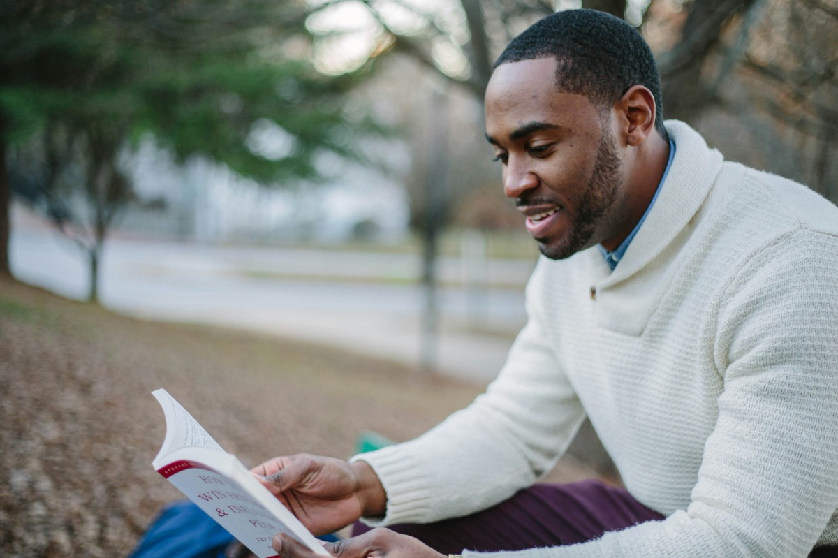 A man in a white sweater sits outdoors on a bench, reading a book. Trees and a path are visible in the background.