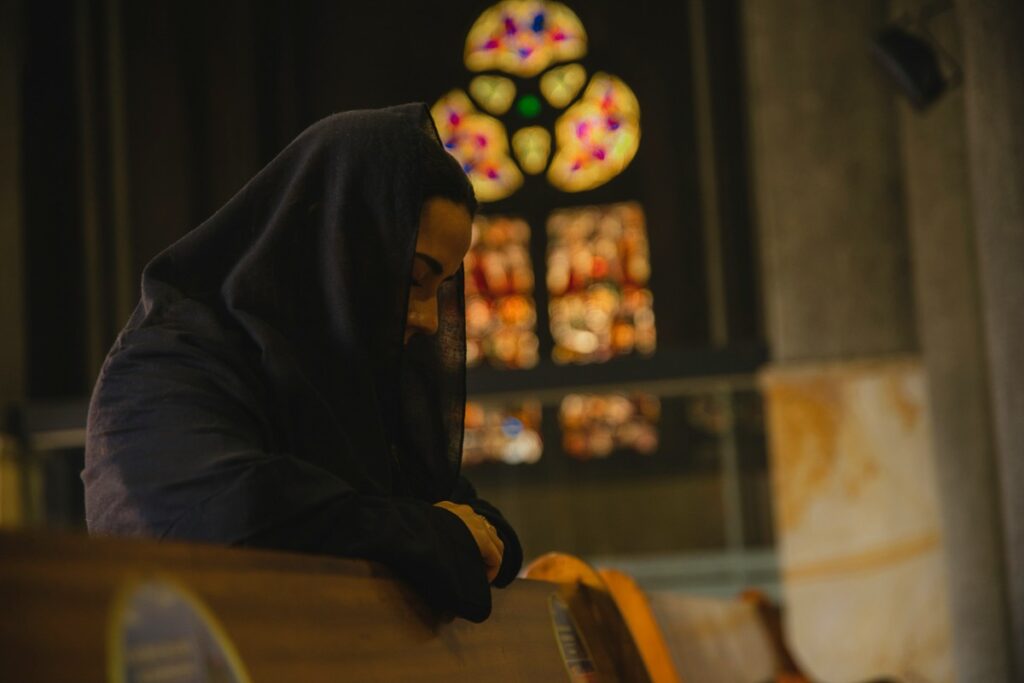 Person in a dark hooded garment praying in a church pew, with a stained glass window in the background.
