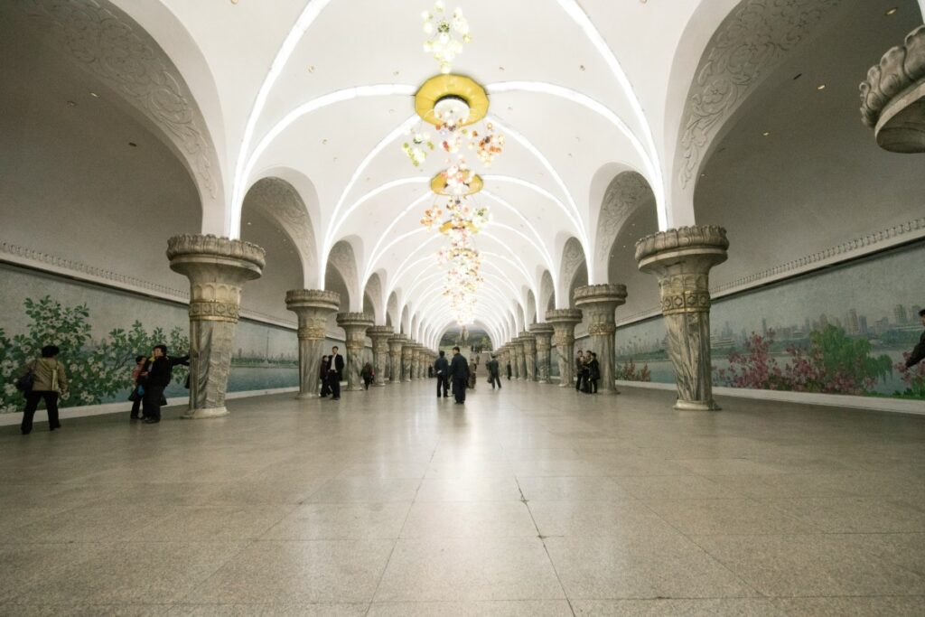 Elegant underground subway station with arched ceilings, ornate chandeliers, and murals on the side walls. Few people walking through the spacious, well-lit hall.
