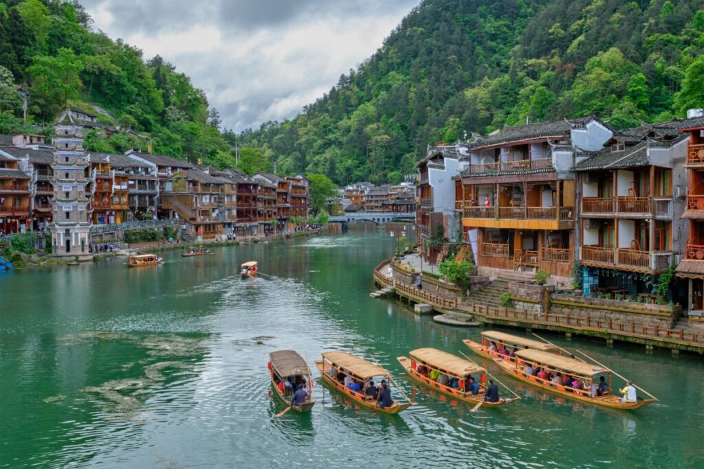 Boats on a river surrounded by traditional wooden buildings and lush green hills under a cloudy sky.
