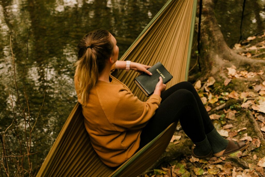 Person sitting in a hammock by a river, holding a notebook, surrounded by fallen leaves and trees.