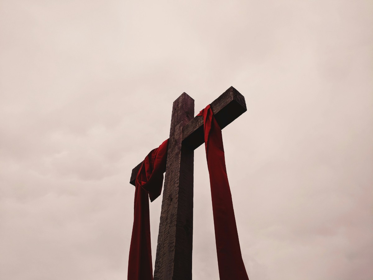 A wooden cross draped with red fabric stands against a cloudy sky.