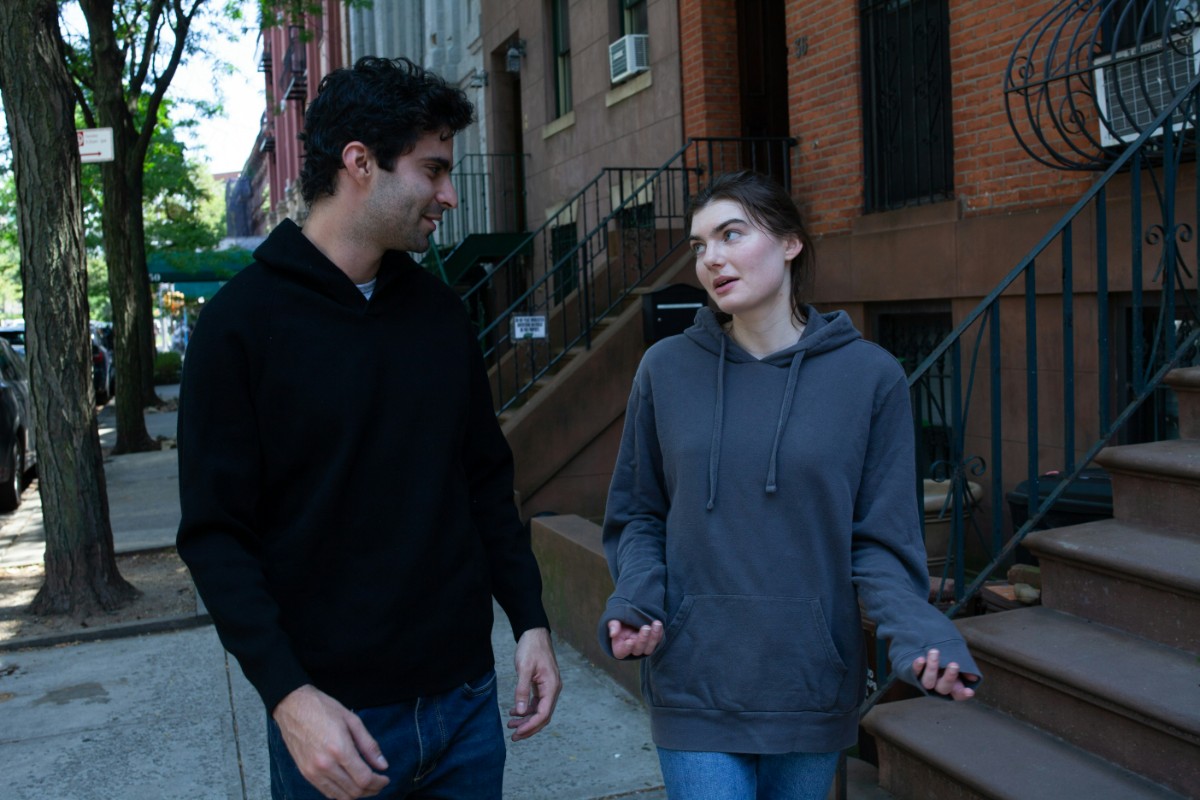 Two people walking and talking on a city sidewalk beside a building with stairs. Both are wearing hoodies, and trees line the street.