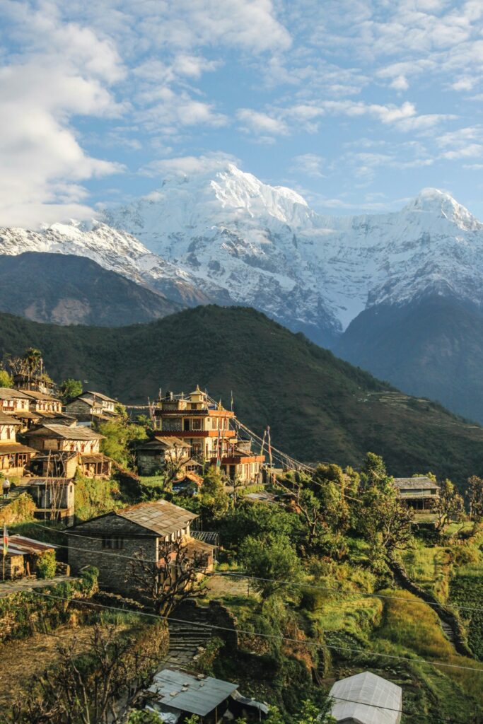 A village with traditional houses nestled among green hills, with a backdrop of snow-capped mountains under a partially cloudy sky.
