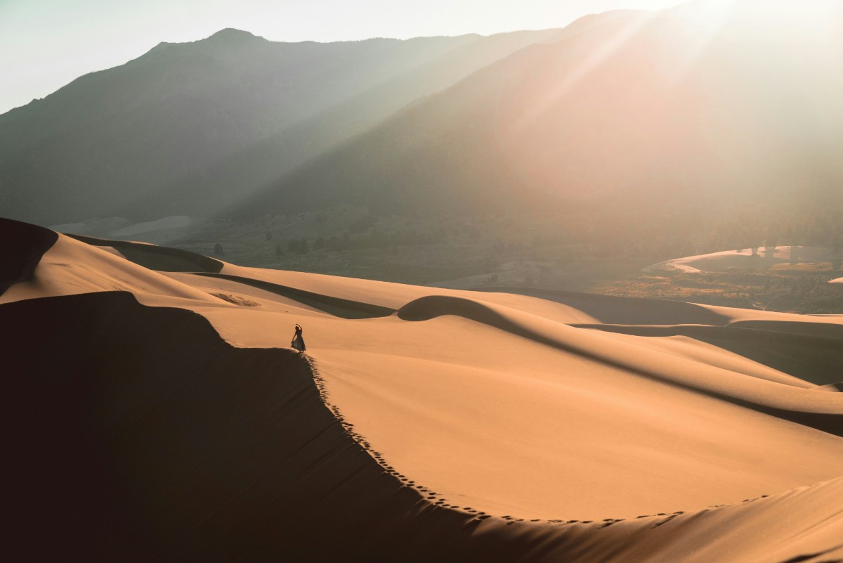 A person walks on sunlit sand dunes with mountainous backdrop, under a bright sky.