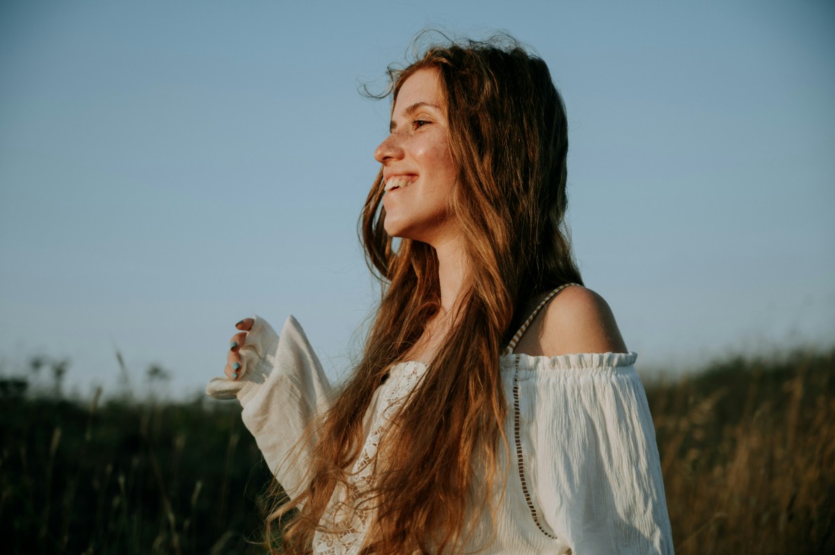 Woman with long hair in a white off-the-shoulder top standing in a field, smiling, with a clear blue sky in the background.