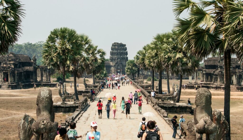 People walking on a wide stone pathway flanked by statues and palm trees, leading towards an ancient temple structure in the background.