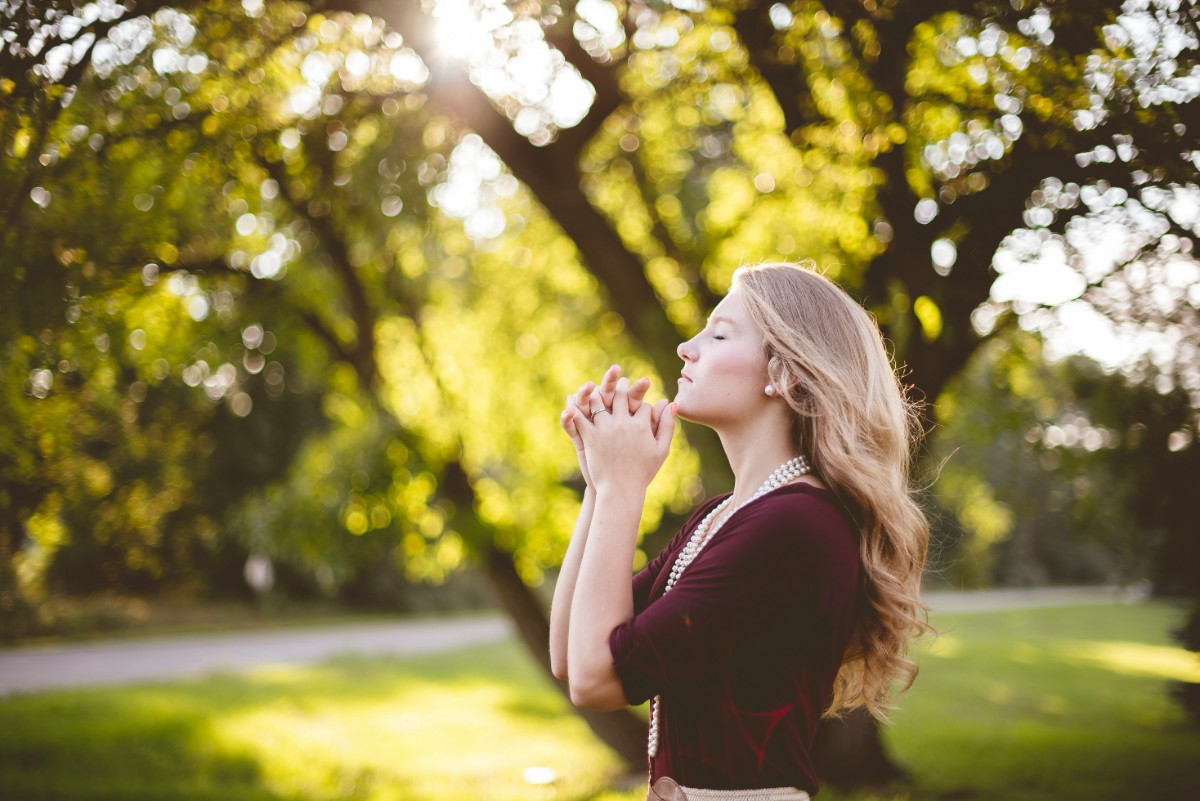 A woman stands outdoors with eyes closed, holding her hands together, surrounded by trees and sunlight filtering through the leaves.