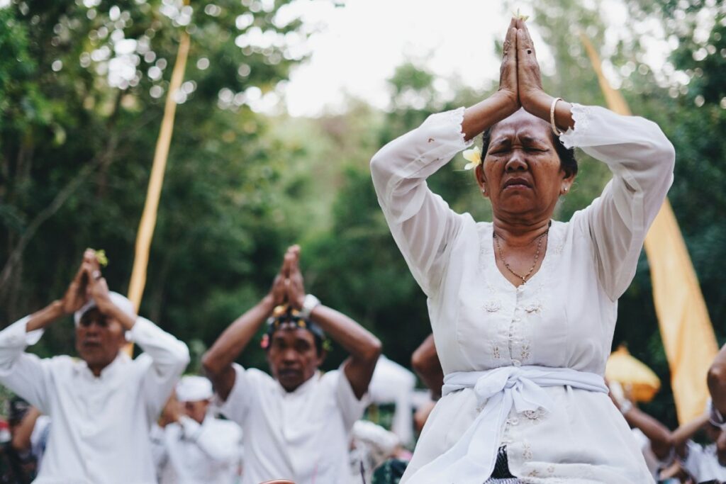 People dressed in white perform a group meditation outdoors, hands pressed together above their heads, surrounded by greenery.