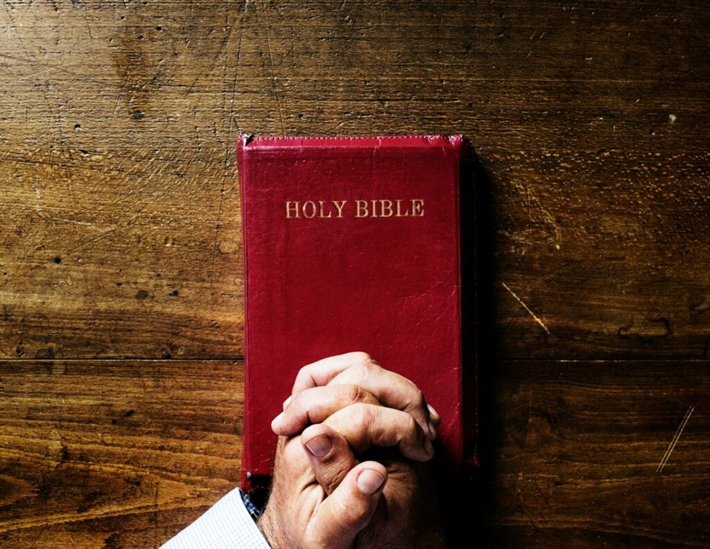 A person with clasped hands rests on a closed red Bible on a wooden table.