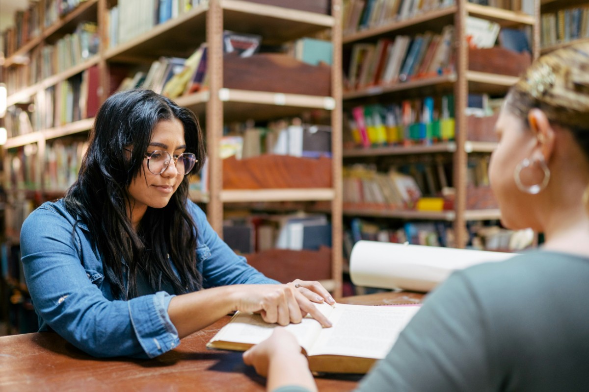 Two people are sitting at a table in a library. One person is pointing at a book they are both looking at. Shelves filled with books are in the background.
