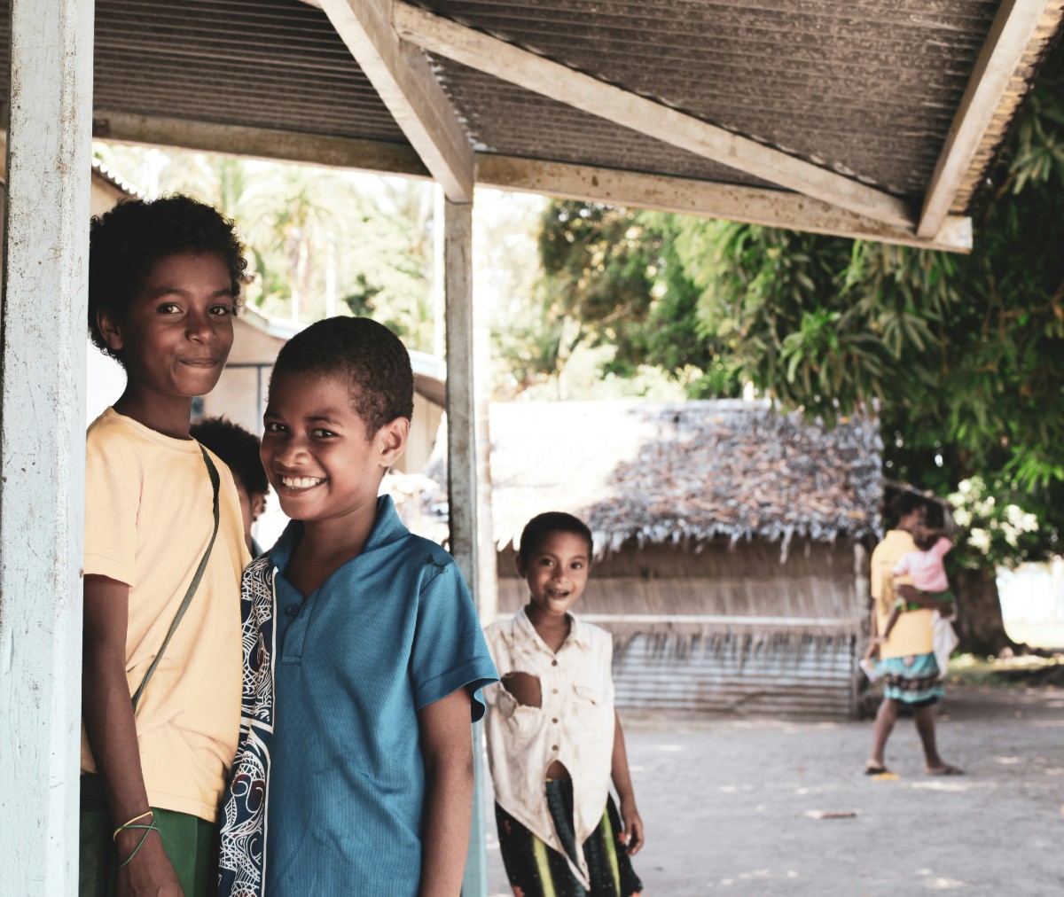 Three children stand under a metal-roofed shelter, with two smiling near a post and one walking in the background. Trees and a building are visible in the background.