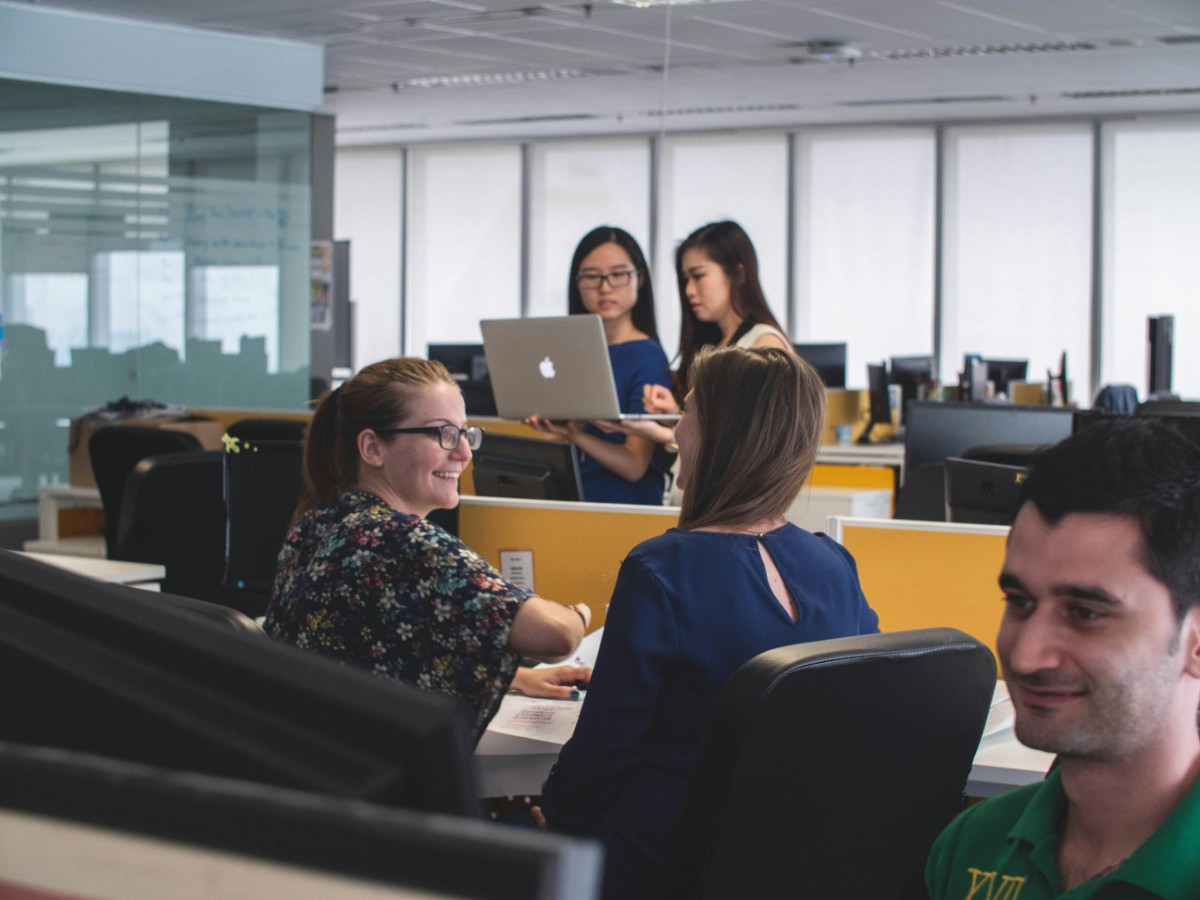 Office environment with five people: two standing and using a laptop, three seated at desks engaged in conversation.