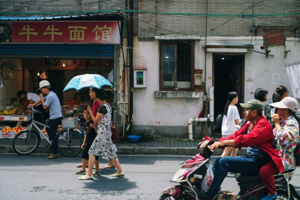 People walk and ride scooters on a street in front of a shop with fruits displayed. A woman holds an umbrella while crossing with a child.