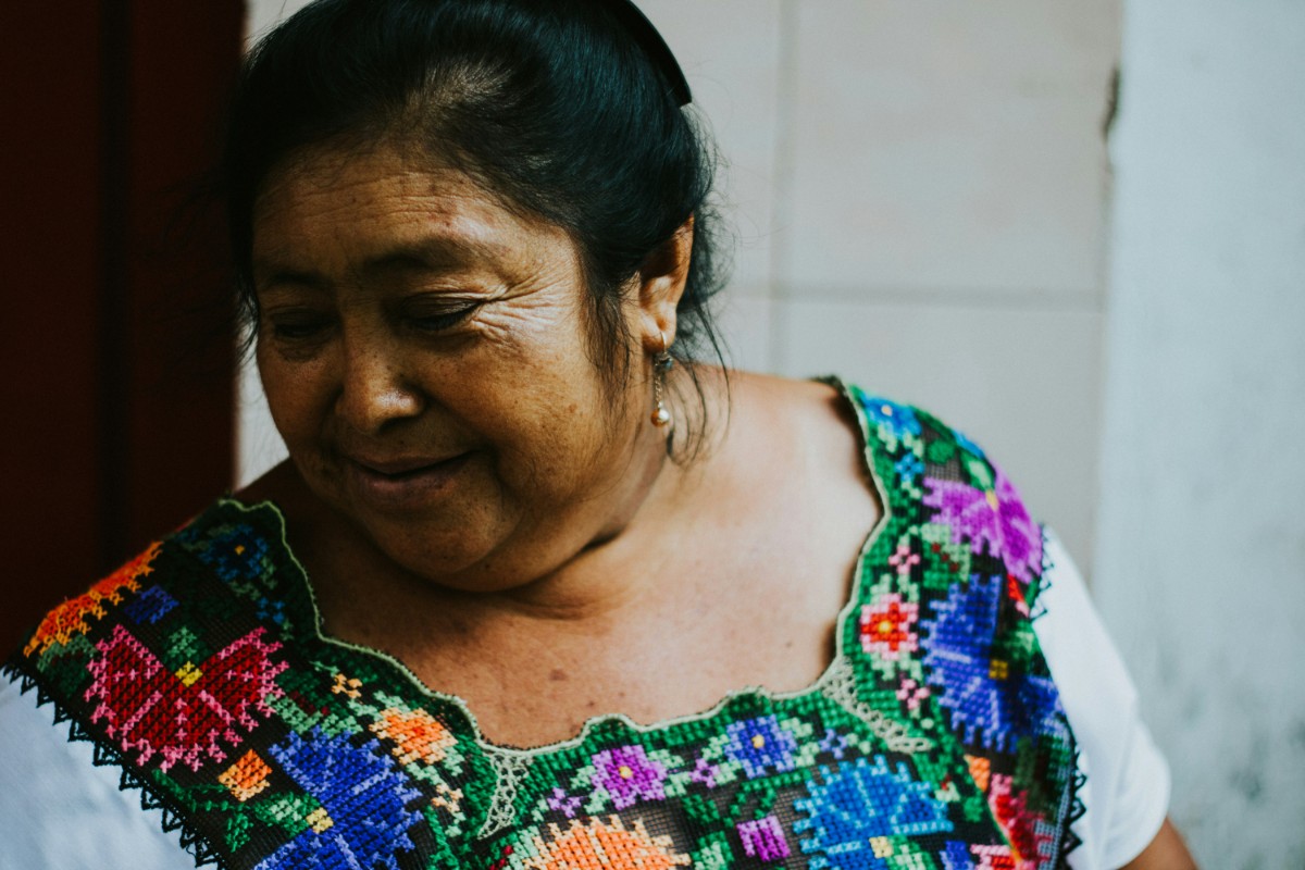 Woman wearing a colorful embroidered blouse, looking down with a neutral expression. Background is a plain tiled wall.