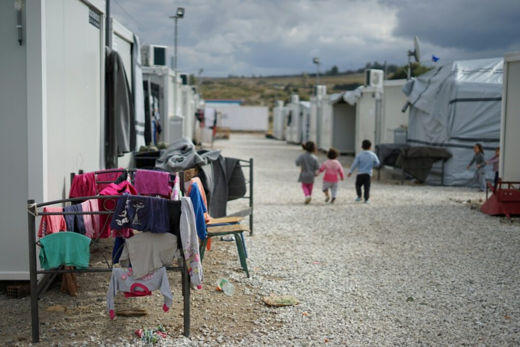 Children walk together in a camp with temporary housing units. Clothes hang on a drying rack in the foreground. Cloudy sky above.