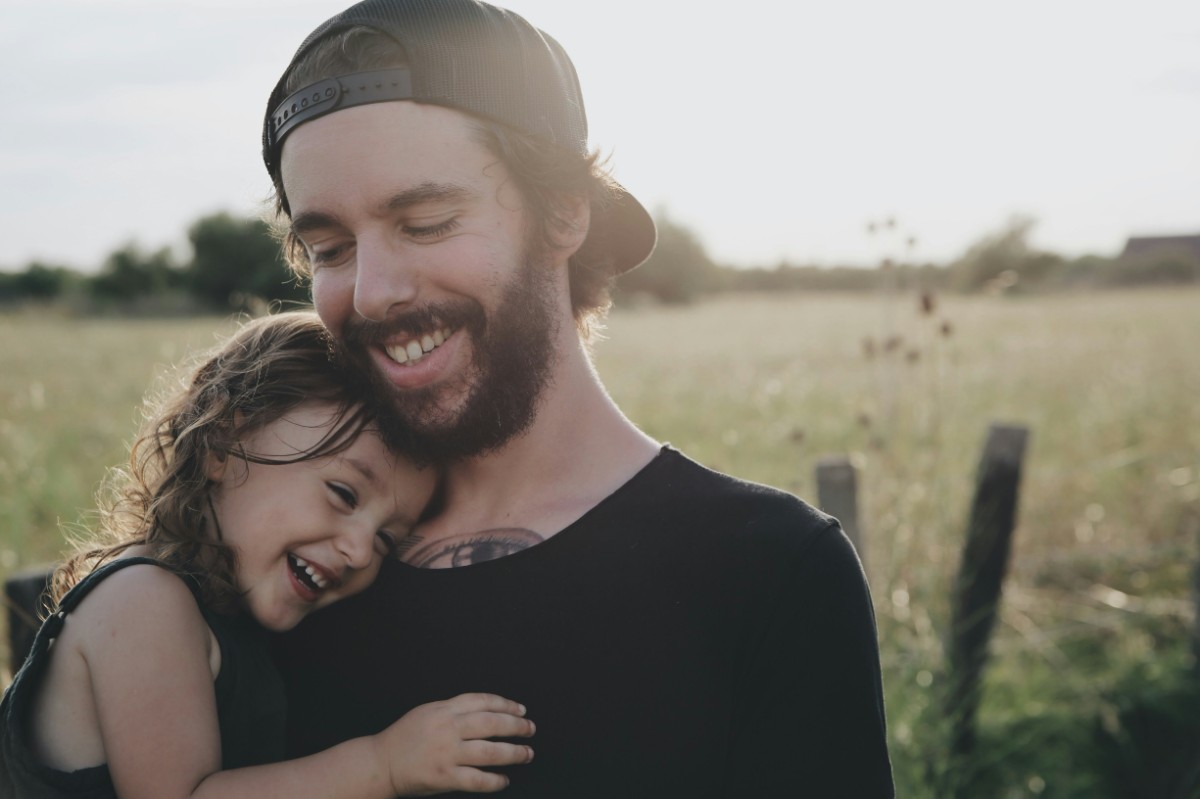 Man and young child smiling and embracing in a grassy field with wooden posts in the background.