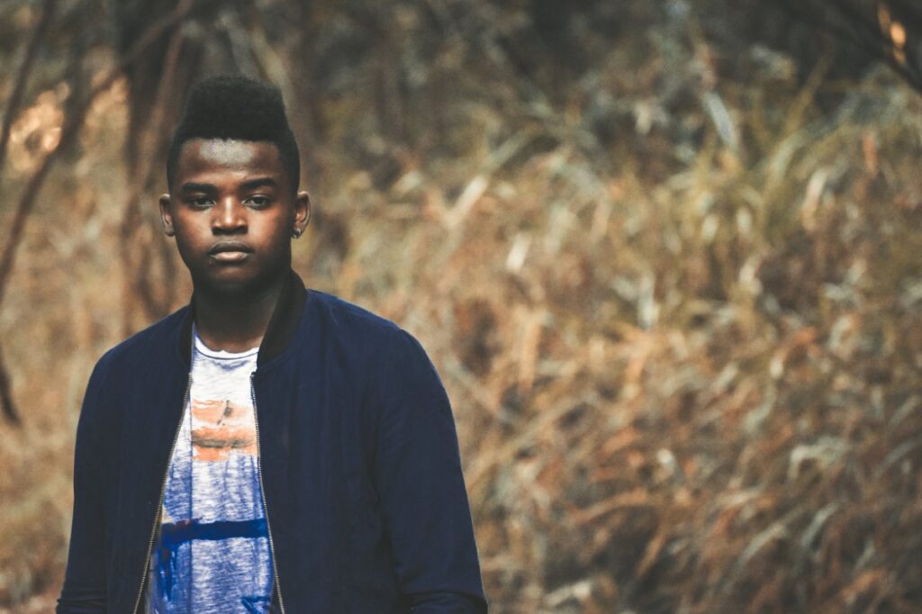 A person stands outdoors in front of dry, tall grass, wearing a dark jacket and a white shirt.