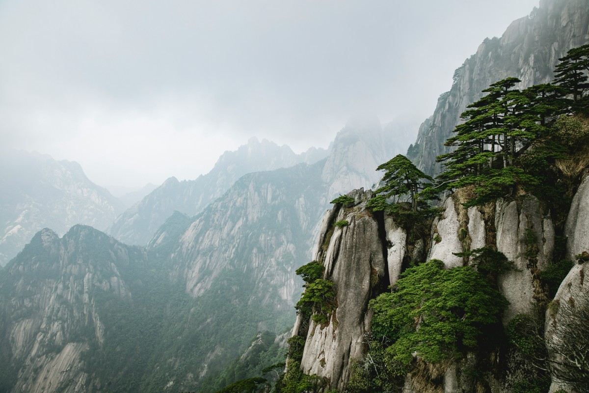 Mist-covered mountain range with steep, rocky cliffs and lush green trees. Cloudy sky enhances the dramatic landscape.