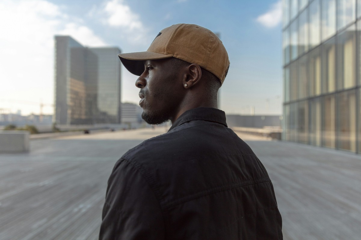 A man in a brown cap and black jacket stands outdoors, looking into the distance. Modern buildings and a partly cloudy sky are in the background.