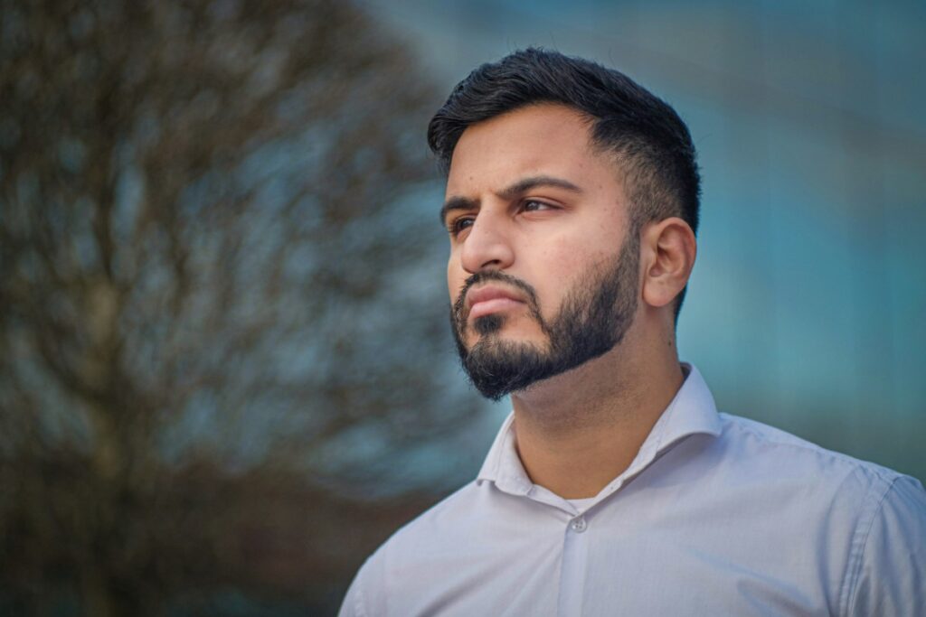 Man in a light shirt looking into the distance with a thoughtful expression, standing outdoors with a blurred tree and building in the background.