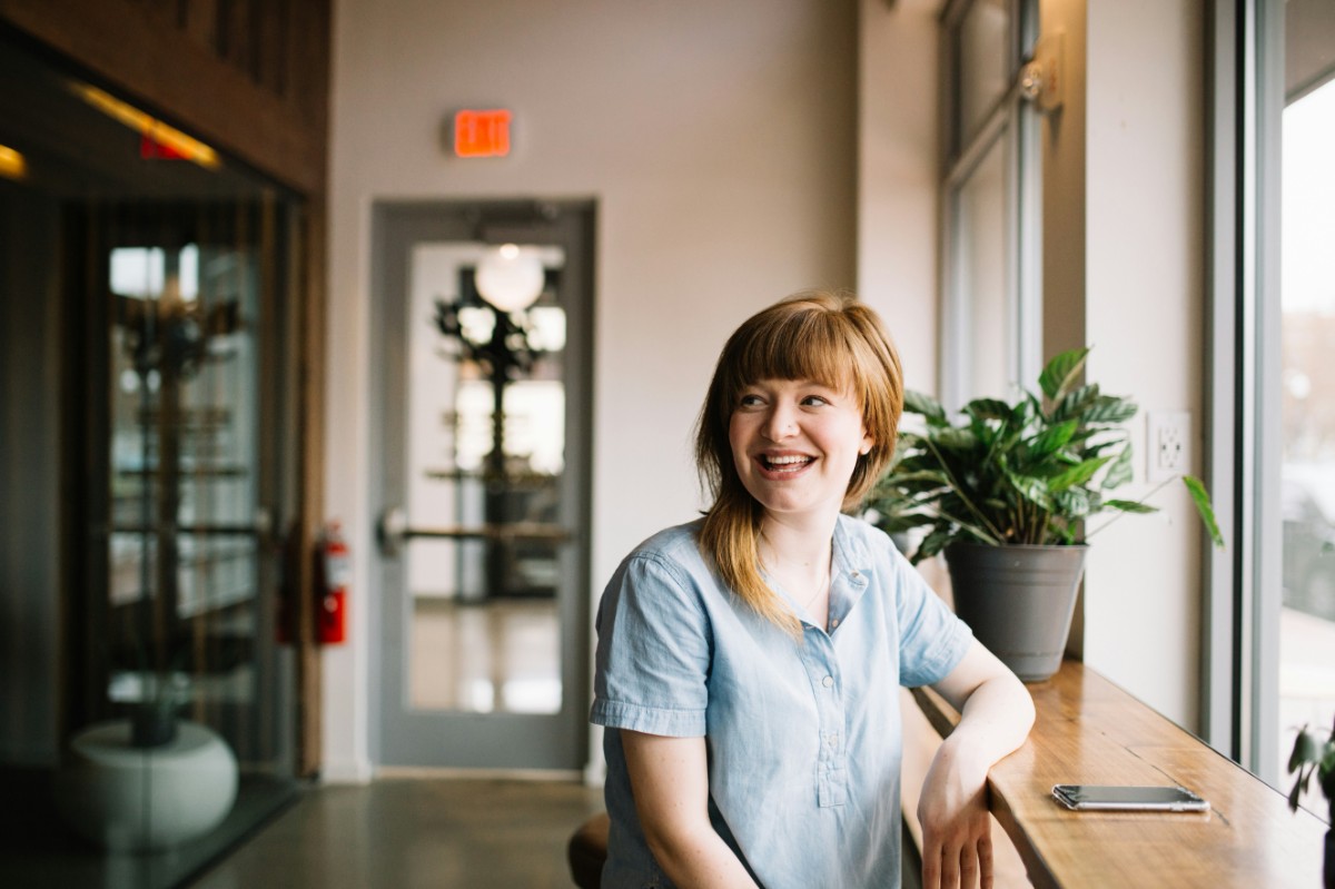 Woman with red hair sits by a window, smiling, in a modern office space. A smartphone and potted plant are on the wooden ledge beside her.