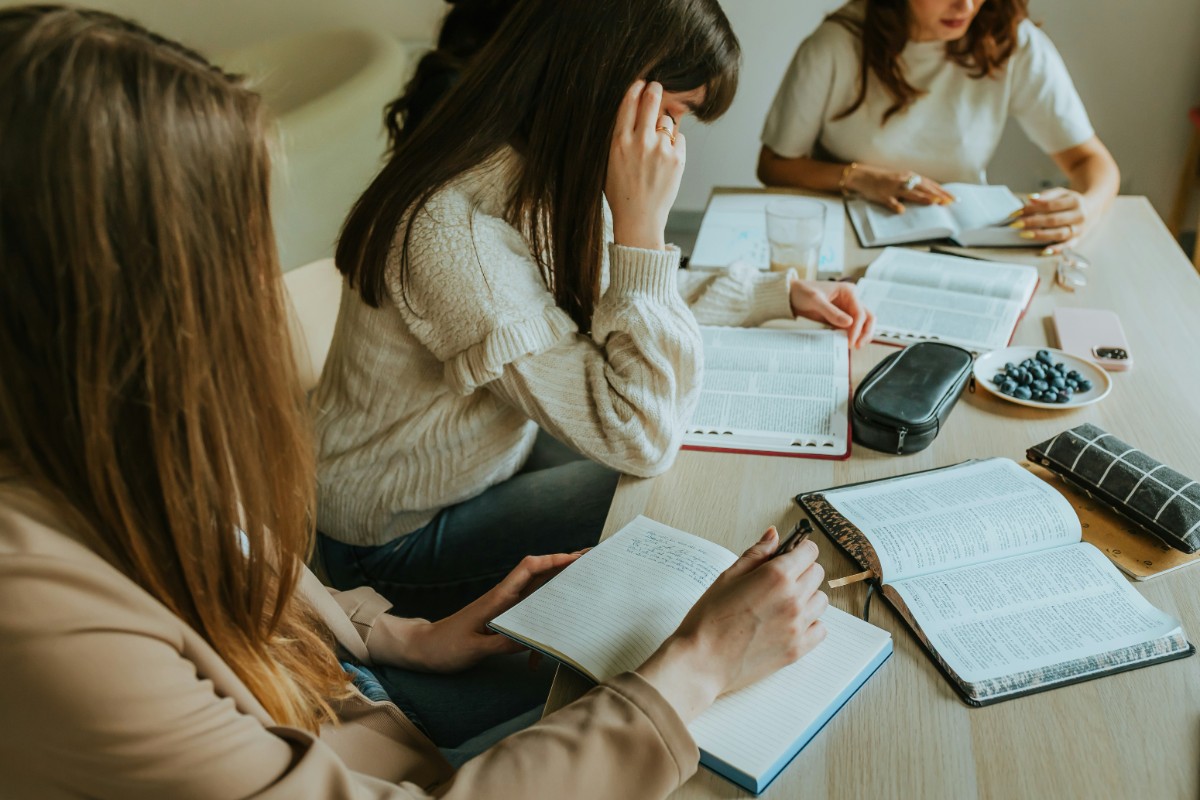 Four people sit at a table, reading and taking notes with books and stationery scattered around.