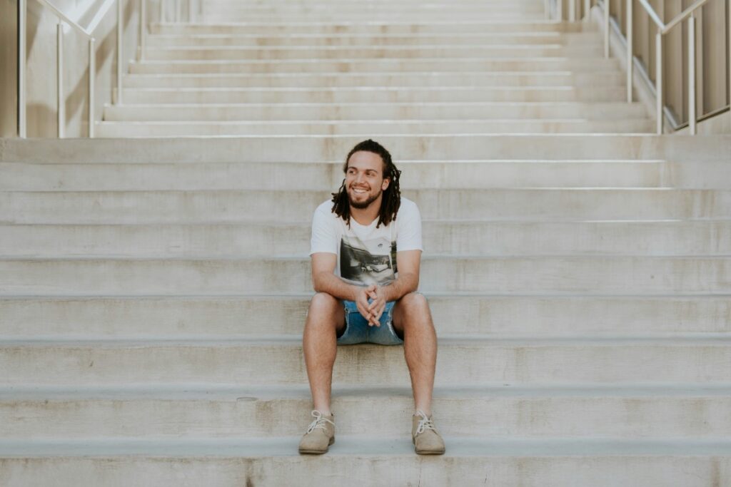A person with long hair sits casually on wide concrete steps, wearing a t-shirt and shorts, looking off to the side with a relaxed expression.