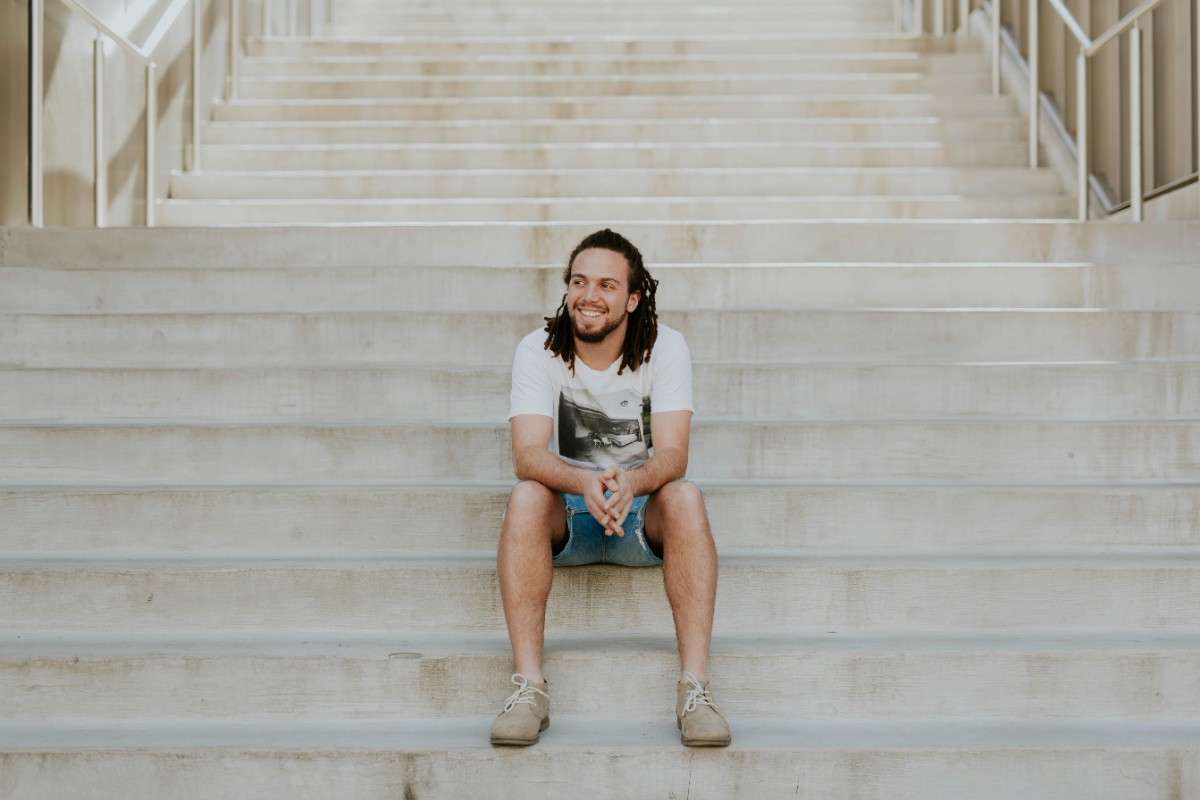 A person with long hair sits casually on wide concrete steps, wearing a t-shirt and shorts, looking off to the side with a relaxed expression.