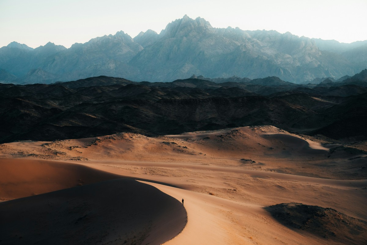 A lone person walks on a sandy desert path with rugged mountains in the background under a clear sky.