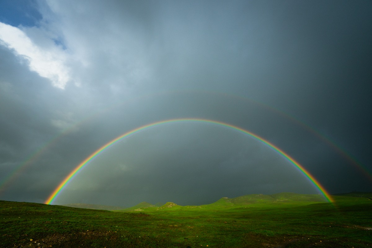 A double rainbow arcs over a green landscape under a cloudy sky.
